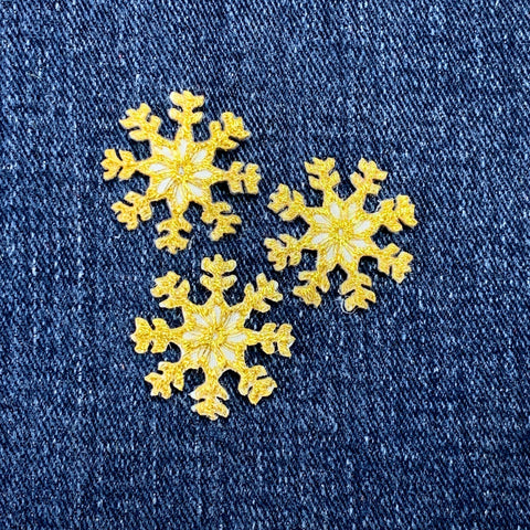 Three small, embroidered, yellow-gold snowflake patches are shown resting on a  denim fabric.