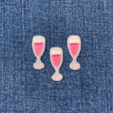 Three embroidered patches shaped like champagne glasses filled with a pink liquid, displayed on a denim background