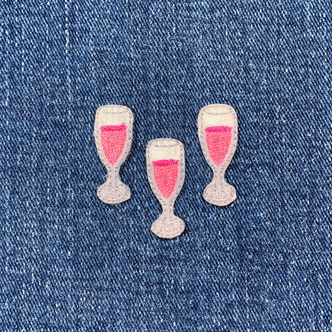 Three embroidered patches shaped like champagne glasses filled with a pink liquid, displayed on a denim background