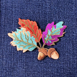 A colorful embroidered patch depicting autumn leaves and acorns on a denim background