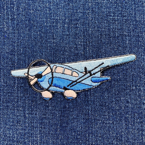 A blue and gray embroidered airplane patch with an iron on backing, displayed on a denim background
