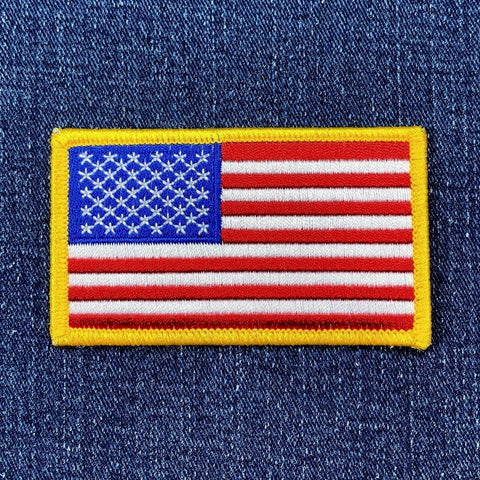 A patch with the design of the United States of America flag, featuring red and white stripes with a blue field and white stars, bordered by a yellow edge, displayed on a denim background