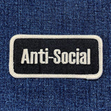 A rectangular black patch with the words 'Anti-Social' in white text, bordered with a white thread displayed on a denim background