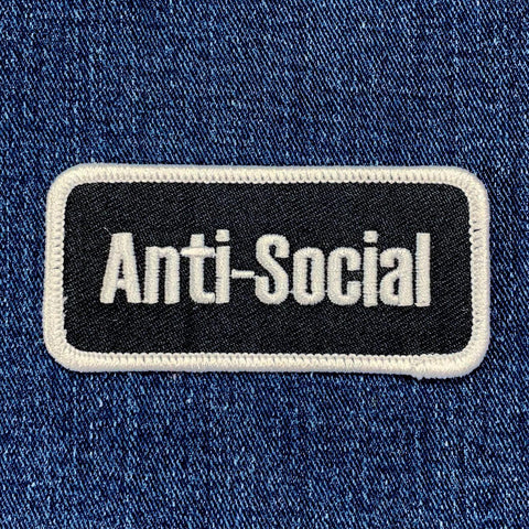 A rectangular black patch with the words 'Anti-Social' in white text, bordered with a white thread displayed on a denim background