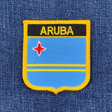 A shield-shaped patch with a blue background, yellow stripes, and the name 'ARUBA' in bold letters, along with a red star, on a denim background