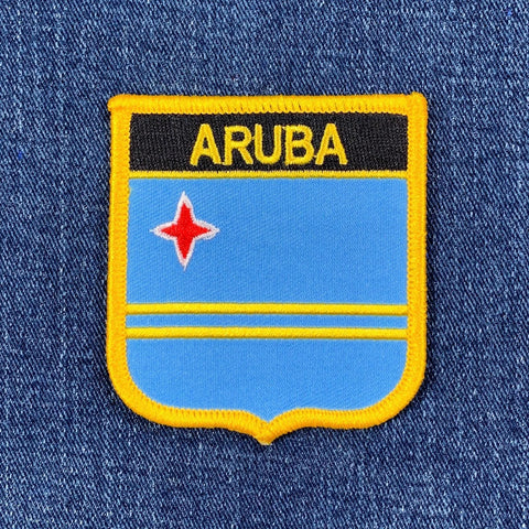 A shield-shaped patch with a blue background, yellow stripes, and the name 'ARUBA' in bold letters, along with a red star, on a denim background