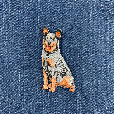 A small embroidered Australian Cattle Dog patch in tan, black, and gray sits on a blue denim fabric background.