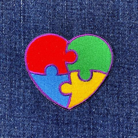 A heart-shaped patch with a puzzle piece design, featuring colors red, blue, green, and yellow, with a purple outline, representing autism awareness, displayed on a denim background