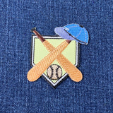 an embroidered baseball patch featuring two baseball batts, a baseball cap, and a baseball positioned over home plate, displayed on a denim background
