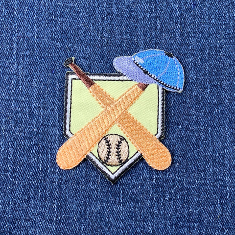 an embroidered baseball patch featuring two baseball batts, a baseball cap, and a baseball positioned over home plate, displayed on a denim background