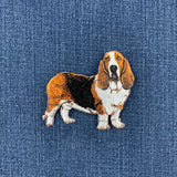 An embroidered basset hound patch in brown, white, and black, placed on a blue denim fabric background.