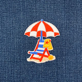 Embroidered patch of a beach scene with an umbrella and chair on denim fabric.