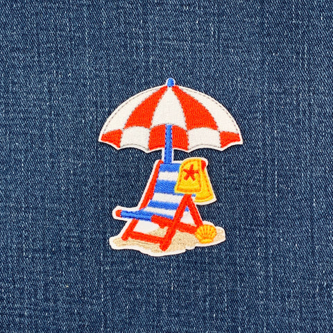 Embroidered patch of a beach scene with an umbrella and chair on denim fabric.