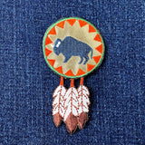A embroidered patch featuring a buffalo round (bison) with feathers around it, displayed on a denim background