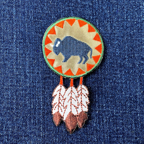 A embroidered patch featuring a buffalo round (bison) with feathers around it, displayed on a denim background