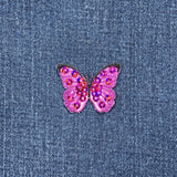 A pink fuchsia butterfly applique patch with mini sequin embellishments and an iron-on backing on a denim background