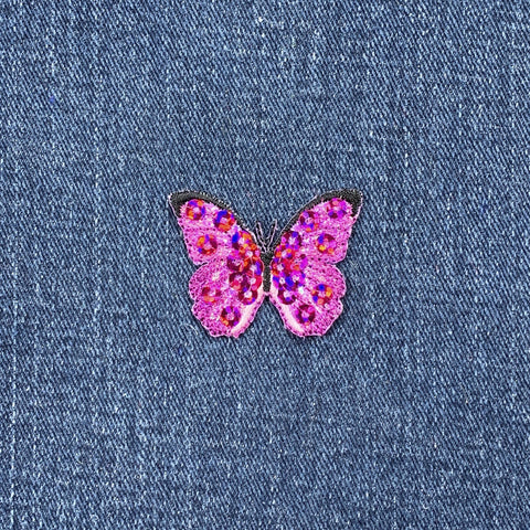 A pink fuchsia butterfly applique patch with mini sequin embellishments and an iron-on backing on a denim background