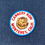 A circular embroidered patch featuring a brown sea otter and the text 'CANNERY ROW MONTEREY, CALIF.' in red on a white background with a blue border, shown on a blue denim surface.