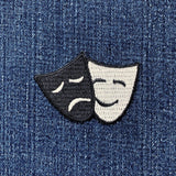 A patch featuring embroidered drama masks, representing comedy and tragedy, designed to be ironed onto fabric, displayed on a denim background