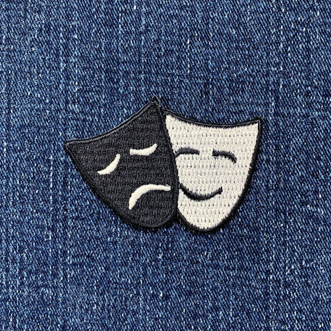 A patch featuring embroidered drama masks, representing comedy and tragedy, designed to be ironed onto fabric, displayed on a denim background