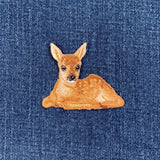An embroidered patch depicting a baby deer, with a realistic design featuring details such as ears, eyes, and a lying pose, displayed on a denim background