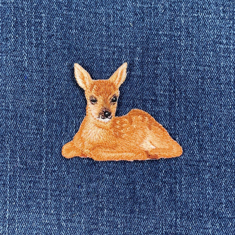 An embroidered patch depicting a baby deer, with a realistic design featuring details such as ears, eyes, and a lying pose, displayed on a denim background