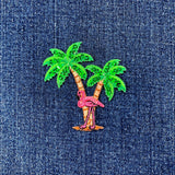 Embroidered patch of a pink flamingo on a palm tree with green leaves on denim fabric.