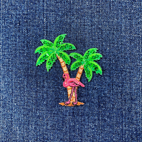 Embroidered patch of a pink flamingo on a palm tree with green leaves on denim fabric.