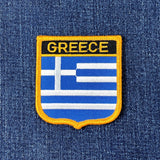 A shield-shaped embroidered patch with the text 'GREECE' and the flag of Greece's stripes in blue and white, displayed on a denim background