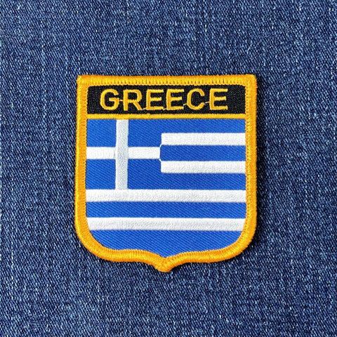 A shield-shaped embroidered patch with the text 'GREECE' and the flag of Greece's stripes in blue and white, displayed on a denim background