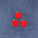 Three embroidered patches shaped like red apples with a bite mark and a green leaf on top on a denim background
