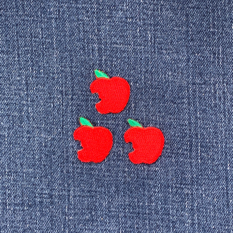 Three embroidered patches shaped like red apples with a bite mark and a green leaf on top on a denim background