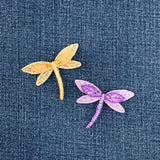 Two embroidered dragonfly patches, one gold and one purple, on a denim background.