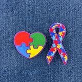 A heart-shaped patch and a ribbon shaped patch with a puzzle piece design, featuring colors red, blue, green, and yellow, with a purple outline, representing autism awareness, displayed on a denim background