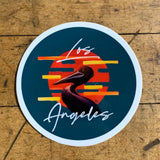 Circular sticker with a dark teal background. The design features a stylized brown pelican silhouette centered over an orange-red circle resembling a sunset, with horizontal yellow and orange stripes. White cursive text above and below the pelican reads "Los Angeles." The sticker is placed on a wooden surface.