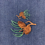 A close-up of a blue denim fabric featuring an embroidered pinecone and pine needles patches in brown and green.