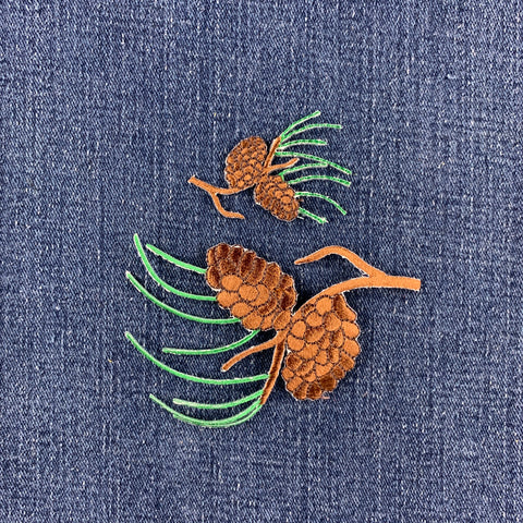 A close-up of a blue denim fabric featuring an embroidered pinecone and pine needles patches in brown and green.