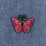 A colorful embroidered butterfly patch with bright pink wings and black antennae, placed on a blue denim fabric background.