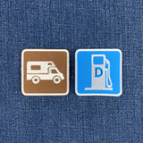 Two square embroidered patches placed on blue denim fabric. The left patch is brown with a white camper van icon. The right patch is a diesel road sign.