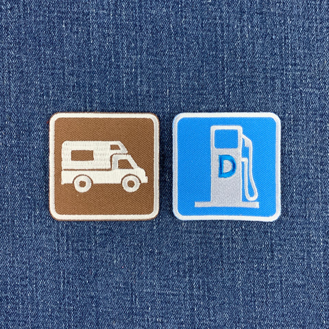 Two square embroidered patches placed on blue denim fabric. The left patch is brown with a white camper van icon. The right patch is a diesel road sign.