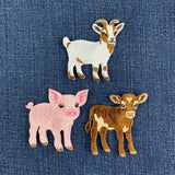 Three embroidered farm animal pins, including a white and brown goat, a pink pig, and a brown and cream calf, displayed on a blue denim background.