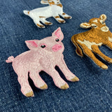 Three embroidered animal patches shaped like a pink pig, a white goat, and a brown cow with cream patches, arranged on a blue denim fabric background.