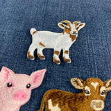 A close-up of embroidered animal patches on blue denim fabric, featuring a white goat with brown patches, a pink pig, and part of a brown-and-white cow.
