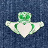 A green and white embroidered patch featuring the Claddagh symbol with a heart and crown on a denim background