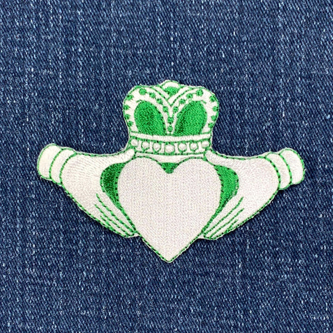 A green and white embroidered patch featuring the Claddagh symbol with a heart and crown on a denim background