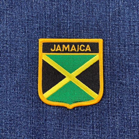 A shield-shaped embroidered patch with the text 'JAMAICA' and the colors of the Jamaican flag on a denim background