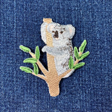 Embroidered patch depicting a koala bear sitting on a eucalyptus tree branch, displayed on a denim background