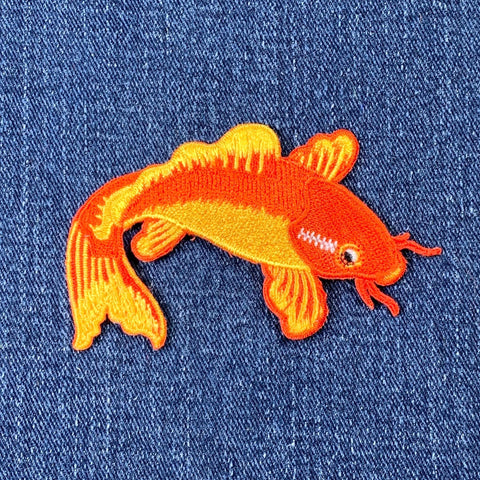 A embroidered patch depicting a koi fish with orange and yellow body on a denim background