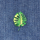 A embroidered applique patch depicting a green Monstera leaf with a tropical pattern on a denim background