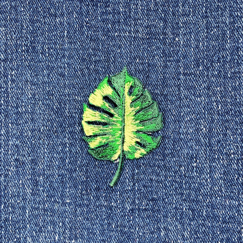 A embroidered applique patch depicting a green Monstera leaf with a tropical pattern on a denim background